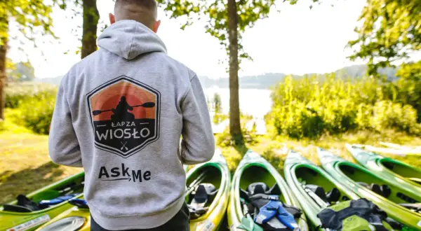Gotowy na spływy kajakowe? Na zdjęciu mężczyzna w bluzie z logo "Łap za wiosło" stoi obok rzędu kajaków nad jeziorem.